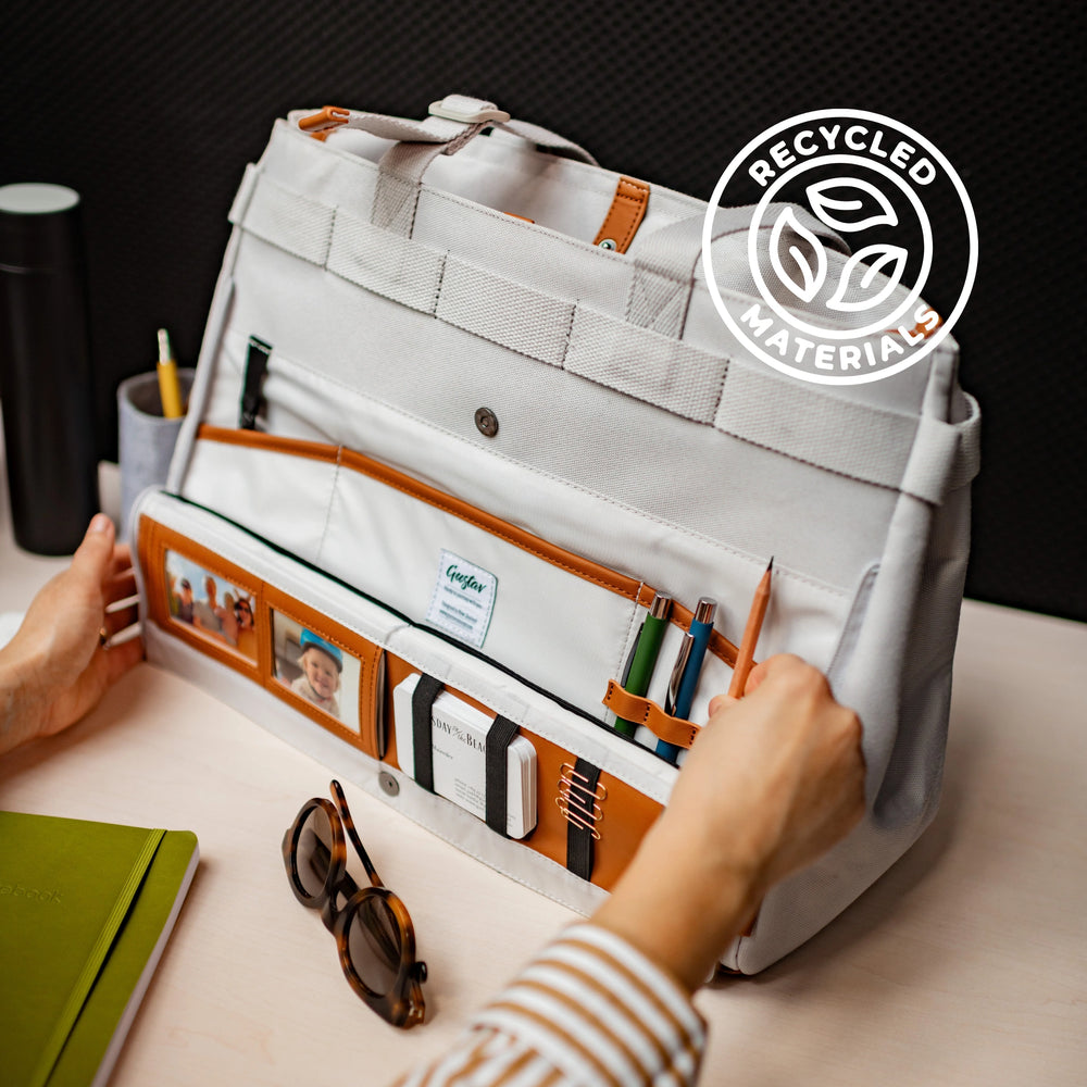 A white and beige DeskMate work bag with brown accents, displayed on a desk, transformed into a workstation with various office supplies like pens, glasses, and a water bottle organized within it.