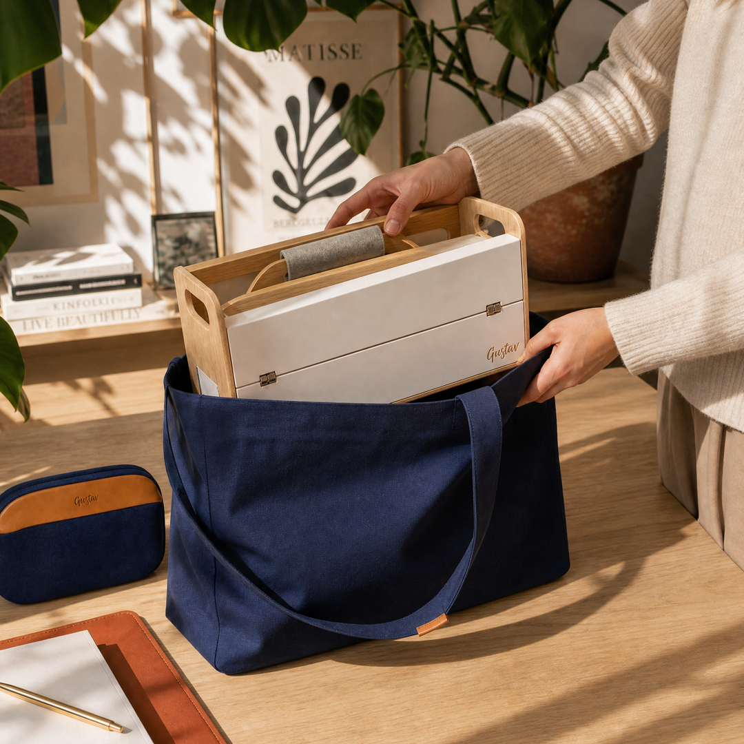 A navy blue canvas tote bag with a wooden handle insert, being held by a person at a wooden desk. A matching navy and tan zippered pouch lies on the desk next to a notebook, with warm sunlight casting leaf shadows across the scene.